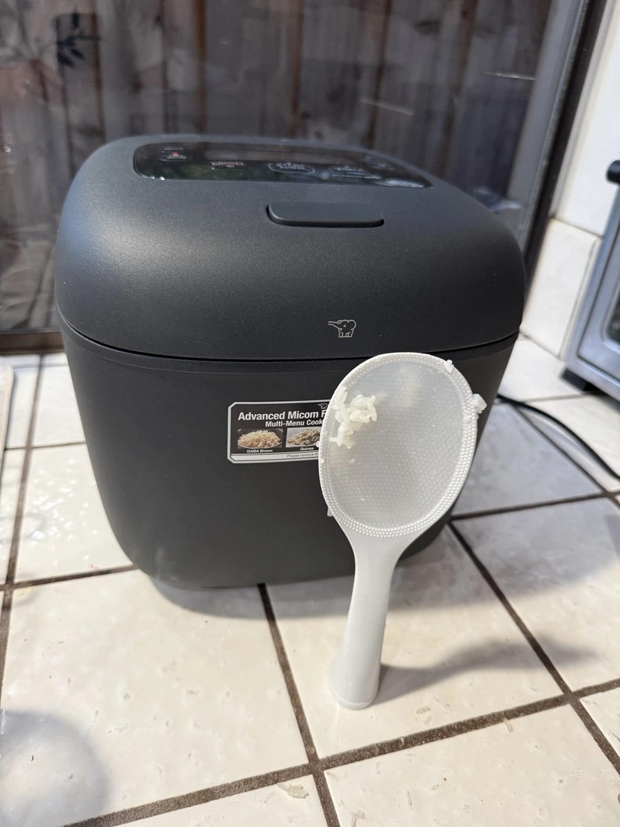 Open Zojirushi NL-EAC18 showing detachable stainless inner lid, standing rice paddle, and measuring cup